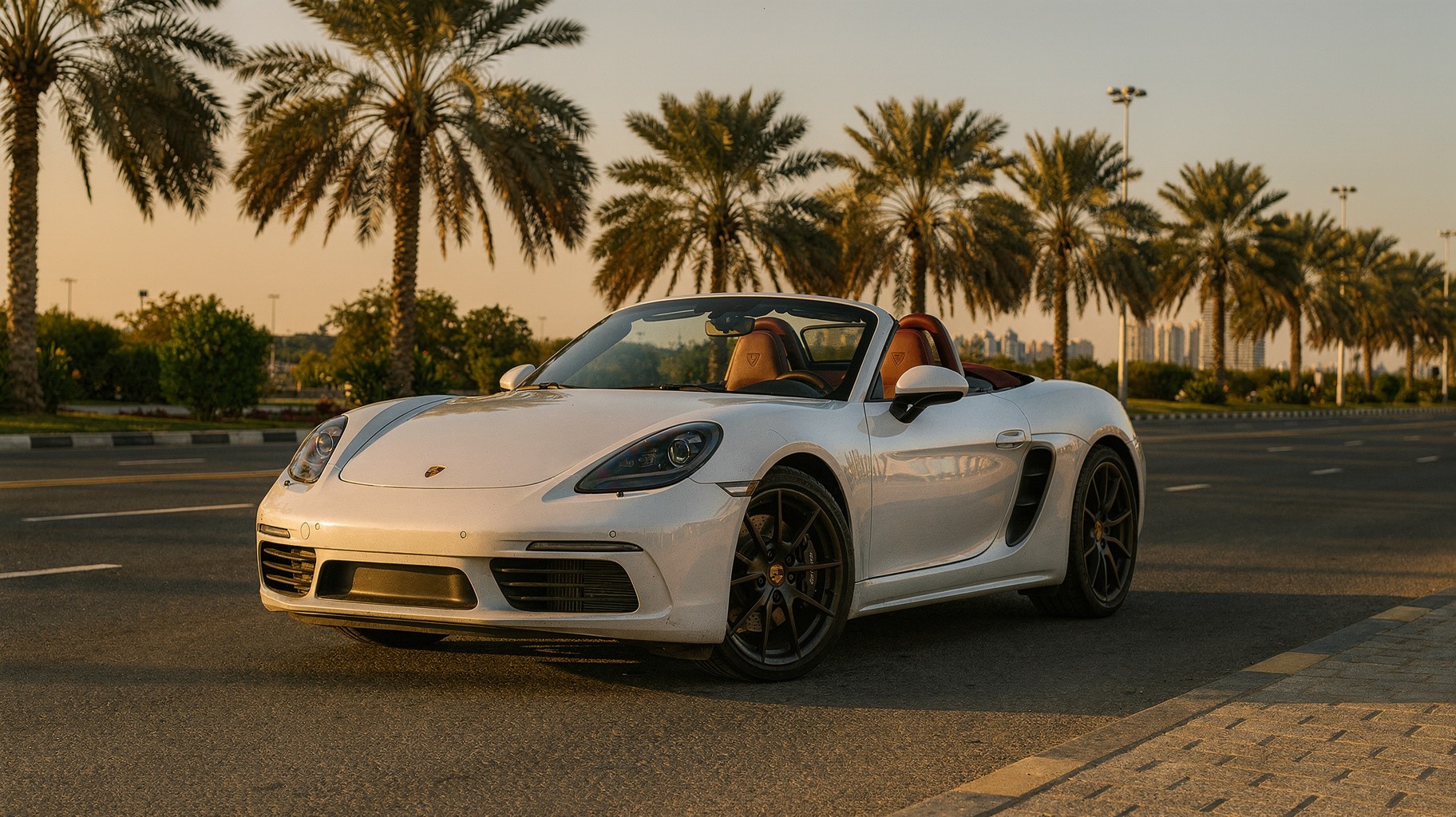 White Porsche 718 Boxster with the roof down on a wide palm-lined Dubai avenue in low winter sunlight.