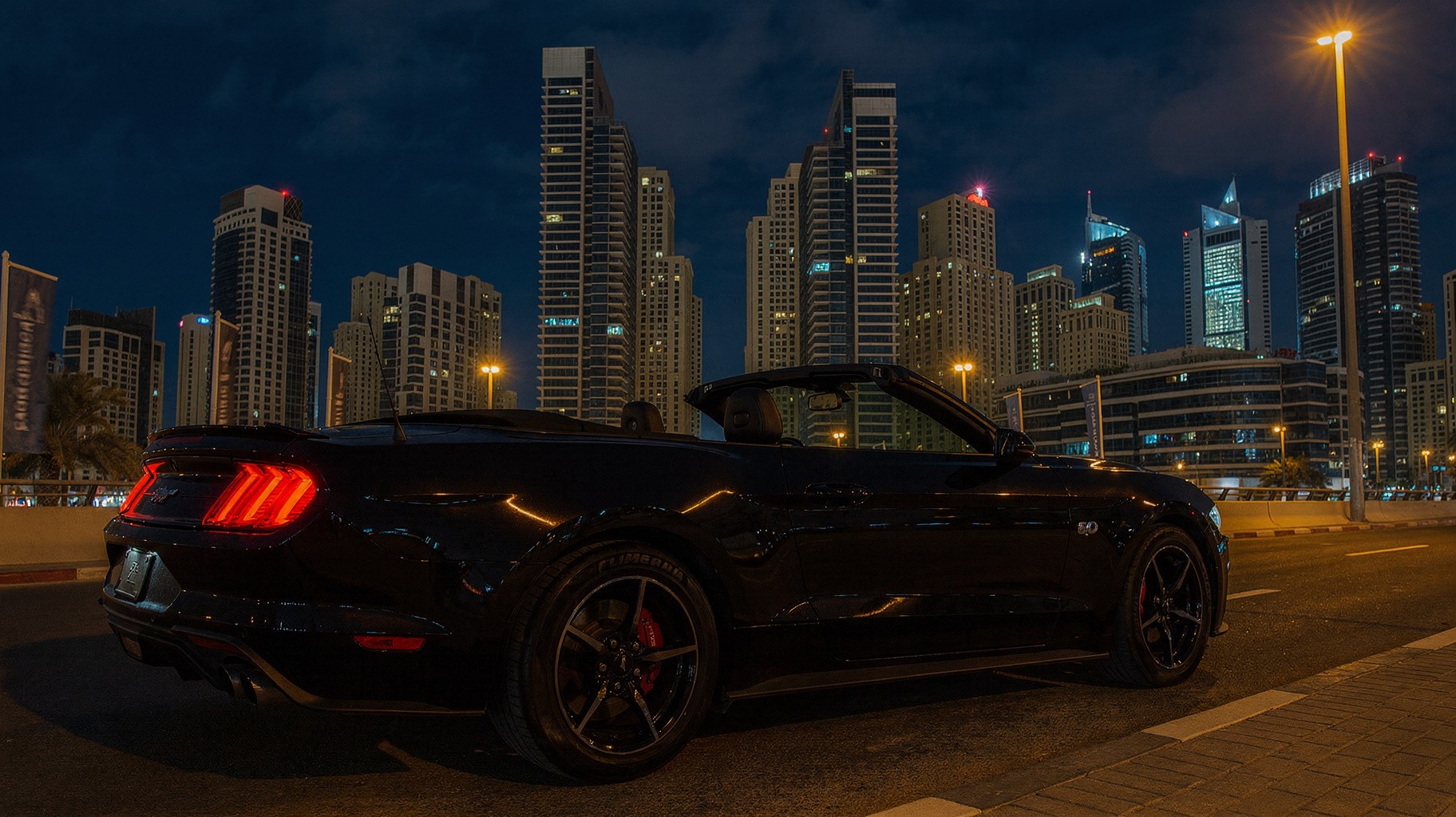 Black Ford Mustang Convertible with the roof down on a Dubai boulevard at night, Marina towers lit behind it.