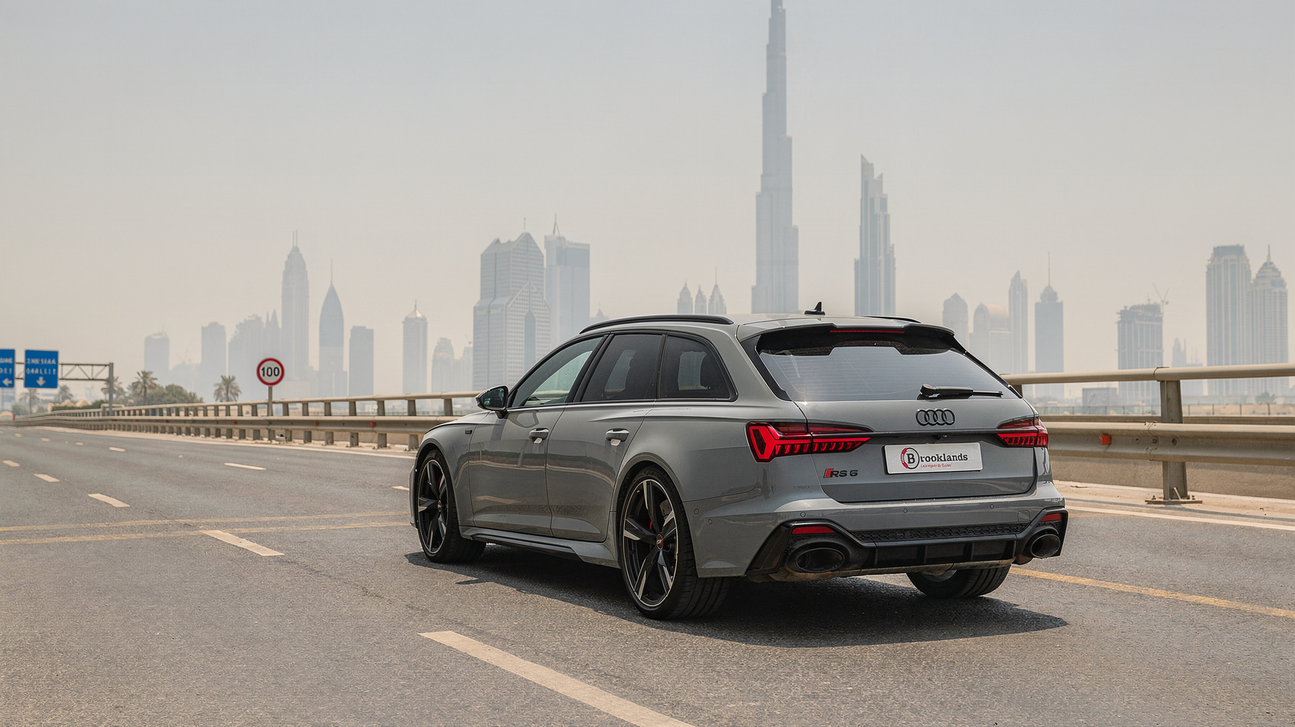 Grey Audi RS6 on an elevated Dubai highway with a 100 km/h speed limit sign and the Burj Khalifa behind it